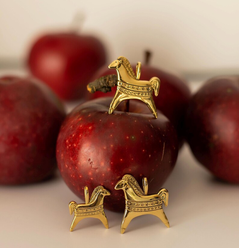Op de afbeelding: Drie goudkleurige paardenbeeldjes, elk met ingewikkelde details, worden getoond op en rond verschillende rode appels. De appels hebben een dieprode kleur en zijn enigszins onscherp op de achtergrond.