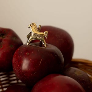 Op de afbeelding: Een gouden paardenbeeldje bovenop een stapel rijpe, rode appels. De appels liggen in een gevlochten mand. Het paard is gedecoreerd met ge&euml;tste patronen. De sc&egrave;ne wordt verlicht door zacht, natuurlijk licht.