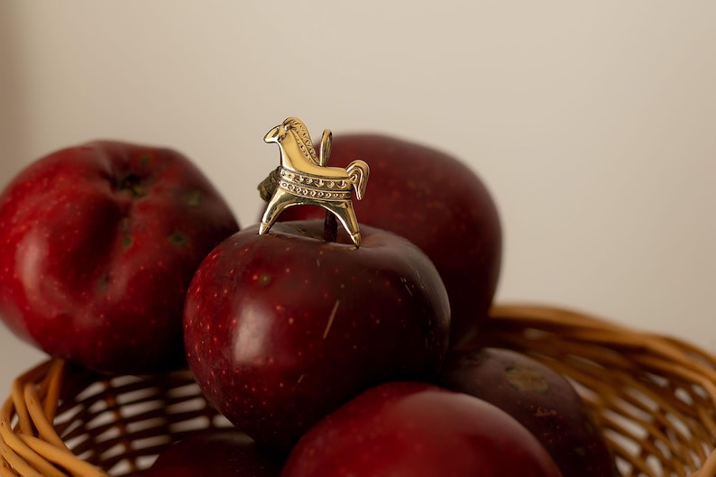 Op de afbeelding: Een gouden paardenbeeldje bovenop een stapel rijpe, rode appels. De appels liggen in een gevlochten mand. Het paard is gedecoreerd met ge&euml;tste patronen. De sc&egrave;ne wordt verlicht door zacht, natuurlijk licht.