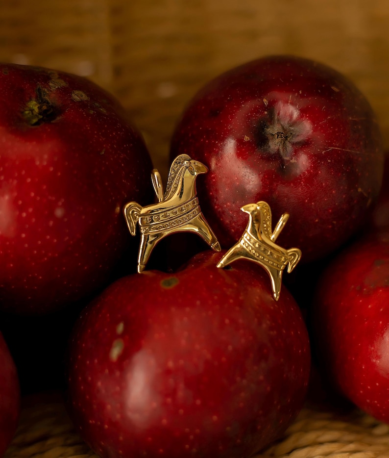 Op de afbeelding: Een paar goudkleurige paardenoorbellen, &eacute;&eacute;n groot en &eacute;&eacute;n klein, liggen op verschillende rode appels. De oorbellen hebben gedetailleerde ontwerpen en staan tegen een vage houten achtergrond.