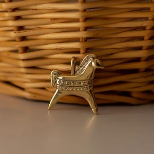 Op de afbeelding: Een goudkleurige paardenhanger met een lus om op te hangen. Het paard heeft een gedetailleerde manen en staart, met decoratieve banden rond het lichaam. De hanger staat tegen een vage achtergrond van een gevlochten mand.