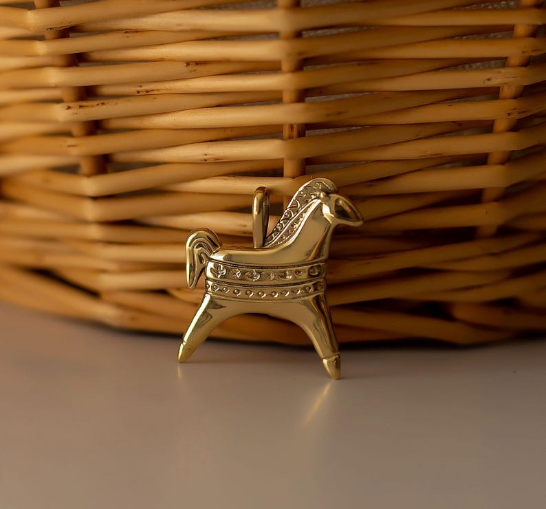 Op de afbeelding: Een goudkleurige paardenhanger met een lus om op te hangen. Het paard heeft een gedetailleerde manen en staart, met decoratieve banden rond het lichaam. De hanger staat tegen een vage achtergrond van een gevlochten mand.