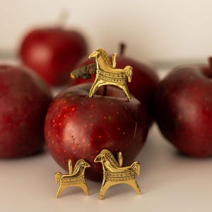 Op de afbeelding: Een stilleven met verschillende rijpe, rode appels en gouden paardenbeeldjes. E&eacute;n beeldje staat op een appel, twee andere ervoor. De appels zijn dieprood, de beeldjes gedetailleerd. De achtergrond is neutraal.