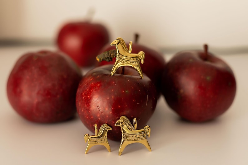 Op de afbeelding: Een stilleven met verschillende rijpe, rode appels en gouden paardenbeeldjes. E&eacute;n beeldje staat op een appel, twee andere ervoor. De appels zijn dieprood, de beeldjes gedetailleerd. De achtergrond is neutraal.