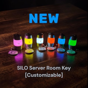 May include: Six cylindrical keychains with glowing, translucent centers in pink, yellow, white, orange, blue, and green. Each features a metallic top and bottom, a numbered center, and a small circular metal charm attached by a silver chain.