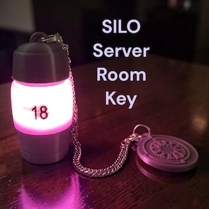 May include: A gray cylindrical keychain with a glowing pink center featuring the number 18, connected by a silver chain to a circular gray medallion with a geometric pattern. The text SILO Server Room Key is visible in the image.