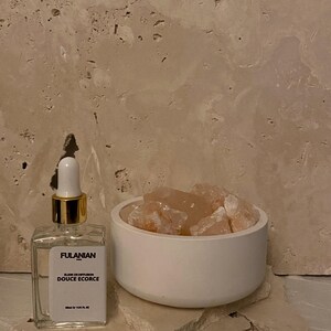 May include: A clear glass bottle with a gold lid and a white label that reads "FULANIAN PARIS OIL OF DIFFUSION DOUCE Ecorce 20ml e 1.02 FL. OZ.". The bottle is sitting on a white surface next to a white bowl filled with pink Himalayan salt rocks.