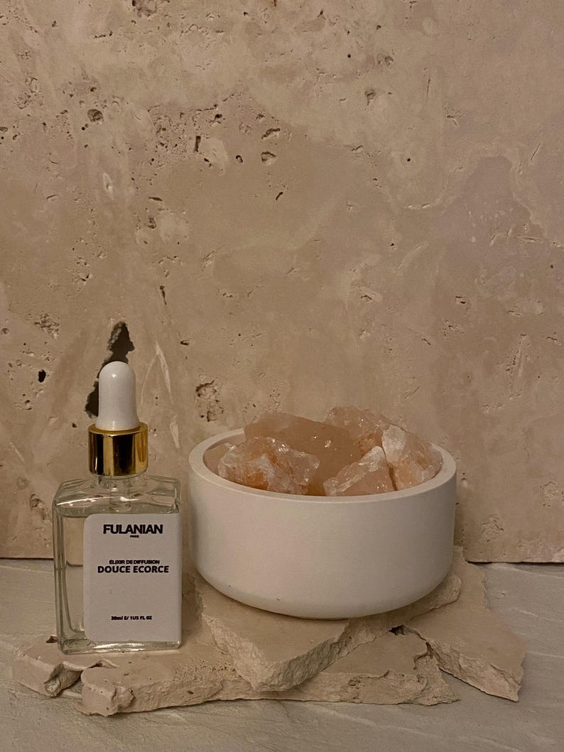 May include: A clear glass bottle with a gold lid and a white label that reads "FULANIAN PARIS OIL OF DIFFUSION DOUCE Ecorce 20ml e 1.02 FL. OZ.". The bottle is sitting on a white surface next to a white bowl filled with pink Himalayan salt rocks.