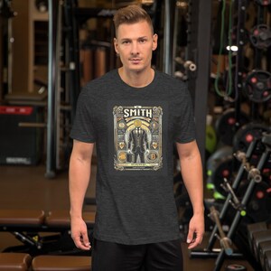 Könnte beinhalten: Dunkelgraues T-Shirt mit detaillierter Grafik. Die Grafik zeigt den Namen "SMITH" oben, eine Anzugfigur mit einem Bitcoin-Symbol für den Kopf, eine Aktentasche und Bitcoin-Symbole. Das Design ist in Gold, Schwarz und Weiß gehalten.