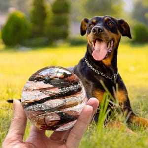 May include: A spherical glass paperweight featuring swirling patterns of black, copper, and white colors with embedded air bubbles. The clear glass orb has a smooth, polished surface and reflects light across its rounded, multi-colored interior design.