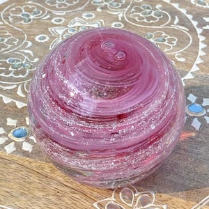 May include: A pink glass orb with white swirls and silver flecks. The orb is sitting on a wooden surface with a decorative pattern.