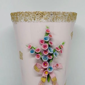 May include: A pink ceramic vase with a gold rim and a floral design. The vase features a bouquet of pink and blue flowers with a pink ribbon.