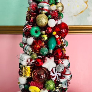 May include: A festive Christmas tree made of ornaments in a white ceramic bowl. The tree is adorned with red, green, gold, and silver ornaments, candy, and lights. The bowl has starburst designs in gold, red, and green.