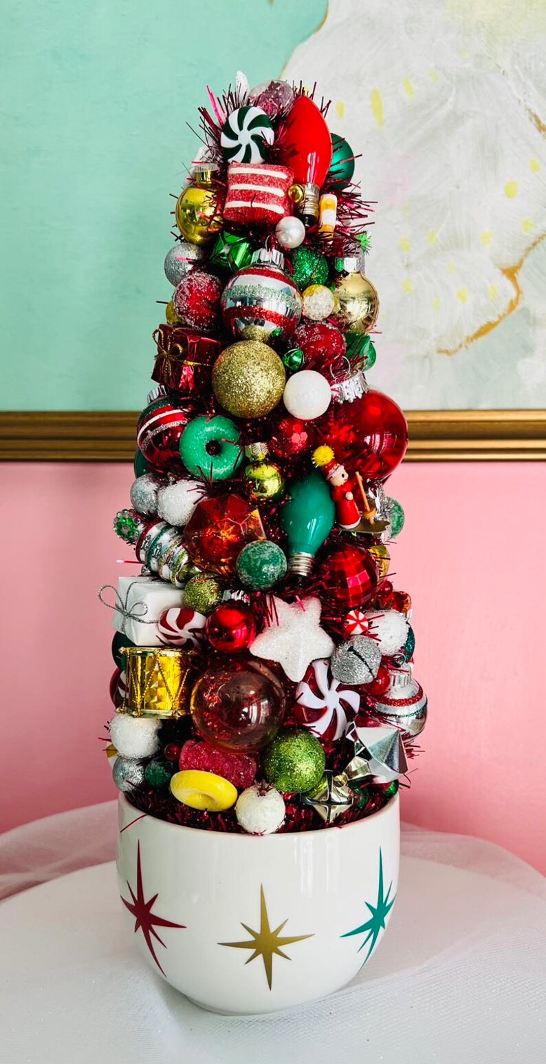 May include: A festive Christmas tree made of ornaments in a white ceramic bowl. The tree is adorned with red, green, gold, and silver ornaments, candy, and lights. The bowl has starburst designs in gold, red, and green.