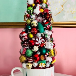 May include: A whimsical Christmas tree made of ornaments, candies, and small decorations, arranged in a white mug with starburst designs. The tree features red, green, gold, and silver ornaments, topped with a white bunny and snowman. A festive holiday decoration.