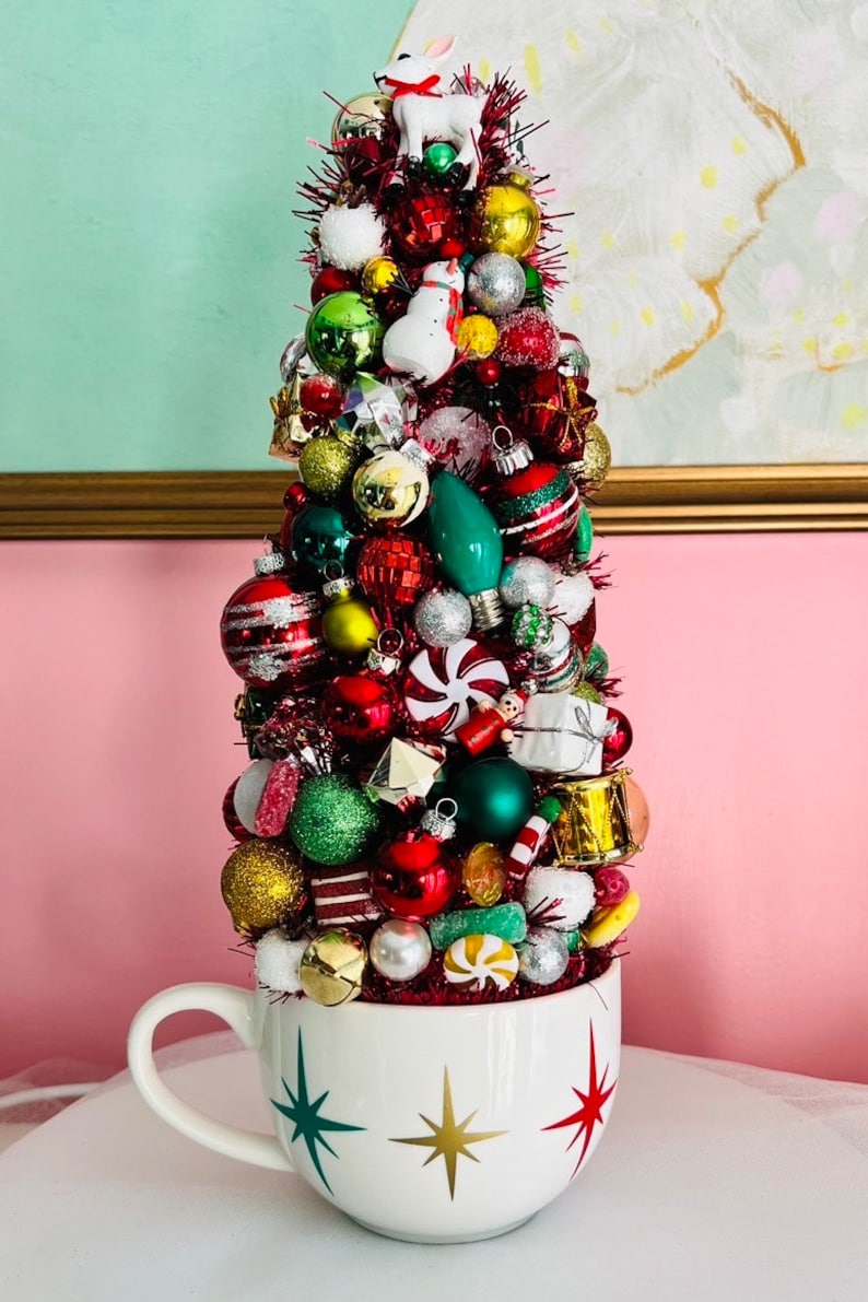 May include: A whimsical Christmas tree made of ornaments, candies, and small decorations, arranged in a white mug with starburst designs. The tree features red, green, gold, and silver ornaments, topped with a white bunny and snowman. A festive holiday decoration.