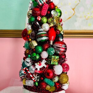 May include: A festive Christmas tree made of various ornaments in a white mug with starburst designs. The tree is adorned with red, green, silver, and gold ornaments, tinsel, and a red lightbulb. A whimsical holiday decoration.