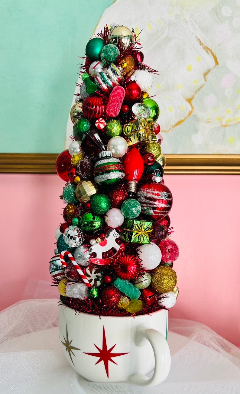 May include: A festive Christmas tree made of various ornaments in a white mug with starburst designs. The tree is adorned with red, green, silver, and gold ornaments, tinsel, and a red lightbulb. A whimsical holiday decoration.