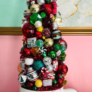 May include: A festive Christmas tree made of various ornaments in a white mug. The tree is adorned with red, green, silver, and gold baubles, candy canes, and small gift boxes. The mug has a starburst design in green, gold, and red.