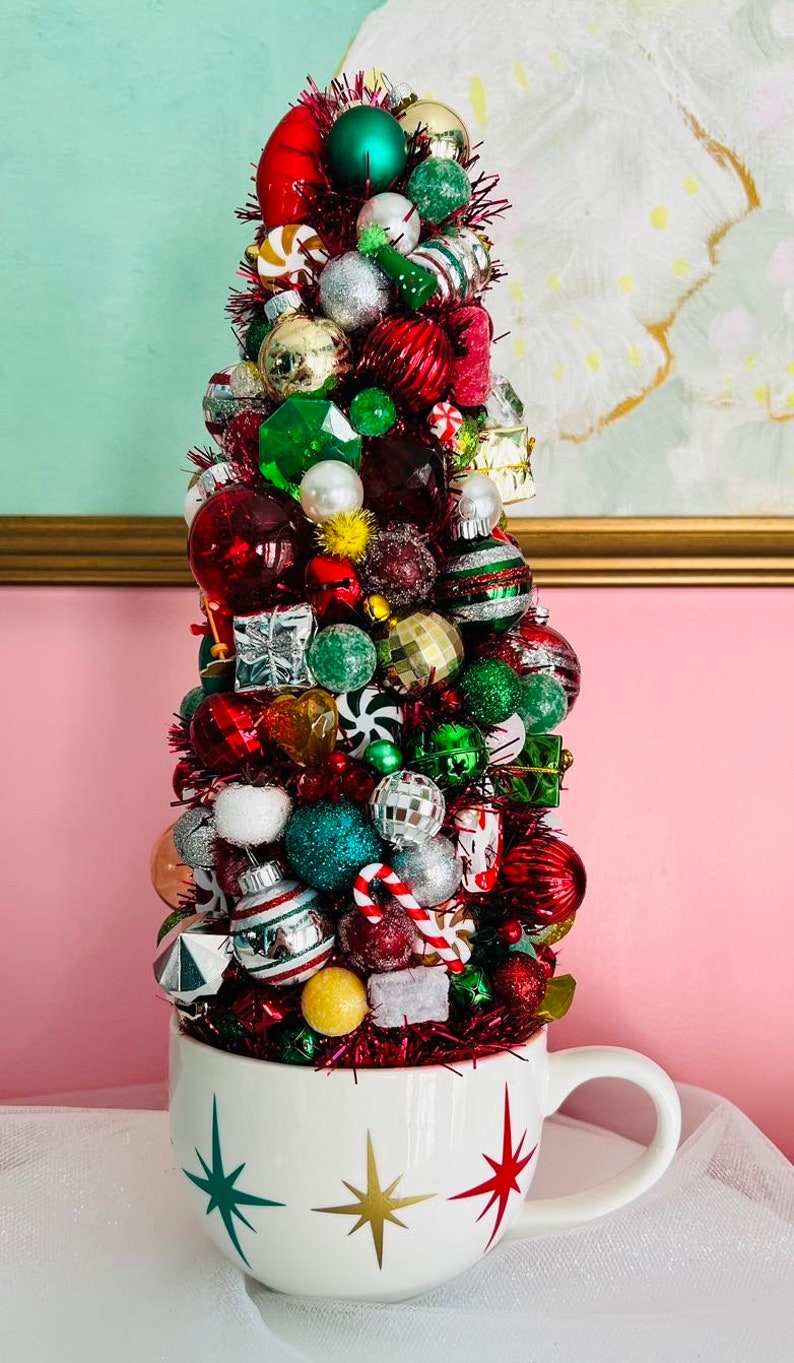 May include: A festive Christmas tree made of various ornaments in a white mug. The tree is adorned with red, green, silver, and gold baubles, candy canes, and small gift boxes. The mug has a starburst design in green, gold, and red.