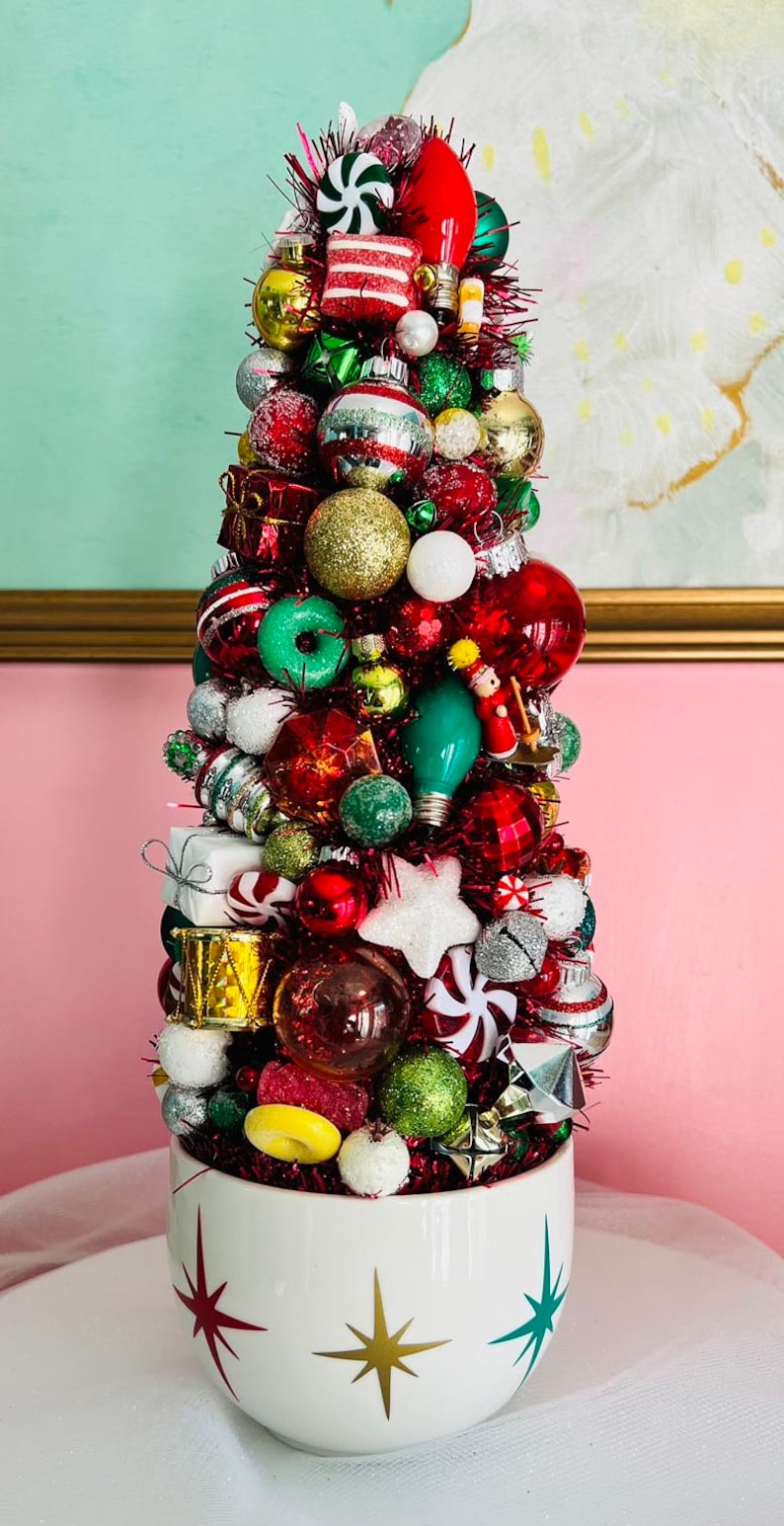 May include: A festive Christmas tree decoration made of various ornaments in red, green, gold, and silver. The tree is in a white ceramic bowl with starburst designs. The ornaments include baubles, lights, and candy.