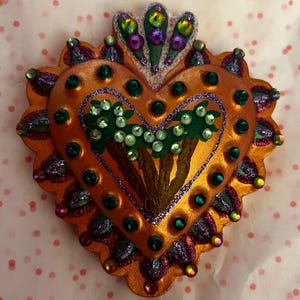 May include: A heart-shaped, orange-colored, decorative object with a raised, three-dimensional design of palm trees. The heart is covered in glitter and rhinestones.