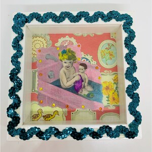 May include: A white frame with a teal sequin border surrounds a vintage illustration of two children in a bathtub. The illustration is on a pink and white patterned paper.
