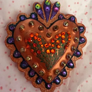 May include: A heart-shaped, copper-colored, decorative object with a scalloped edge. The heart is covered in glitter and rhinestones in shades of orange, purple, and gold. The heart has a painted design of a floral pattern.