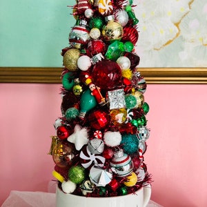 May include: A whimsical Christmas tree made of various ornaments in a white teacup. The tree is adorned with red, green, gold, and silver ornaments, including bulbs, stars, and candy canes. The teacup has a starburst design.