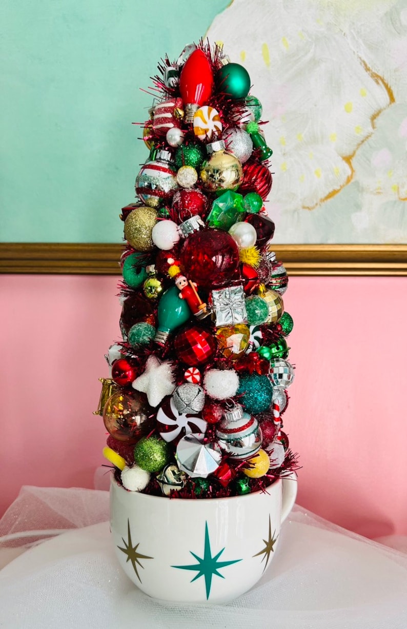 May include: A whimsical Christmas tree made of various ornaments in a white teacup. The tree is adorned with red, green, gold, and silver ornaments, including bulbs, stars, and candy canes. The teacup has a starburst design.