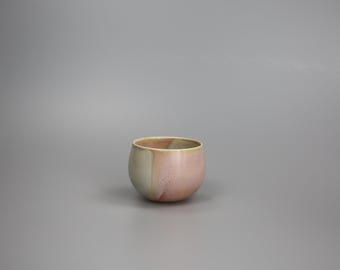 May include: A small, round ceramic bowl with a two-tone color scheme. The bowl is primarily a soft pink, transitioning to a creamy off-white on one side. The rim has a subtle yellow hue. The bowl is likely used for serving or decoration.