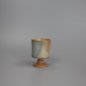 Puede incluir: Un cáliz de cerámica hecho a mano con un cuerpo cilíndrico y una base de pedestal. El cáliz presenta una mezcla de colores, incluyendo azul, beige y marrón. La taza mide aproximadamente 10 cm de alto y es adecuada para bebidas.