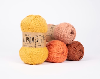 Pode incluir: Uma coleção de novelos de lã coloridos em tons de amarelo, laranja, pêssego e castanho. A lã é rotulada como "Drops Alpaca Uni Colour" com uma ilustração de alpaca. Ideal para projetos de tricô e crochê.