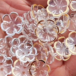 Pode incluir: Close-up de contas transparentes em forma de flor com bordas douradas e prateadas. As contas têm um design facetado, que capta a luz. Estas contas decorativas são ideais para fazer joias e projetos de artesanato.