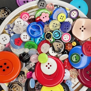 Op de afbeelding: Een verzameling kleurrijke knopen in verschillende maten en vormen. De knopen zijn in tinten rood, blauw, geel, groen, roze, paars, zwart en wit. Sommige knopen hebben vier gaten, andere twee of geen. De knopen zijn gerangschikt in een witte kom.