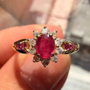 Puede incluir: Un anillo de oro con un gran rubí ovalado central, rodeado de pequeños diamantes y rubíes más pequeños. El anillo tiene un diseño floral con una banda de oro.