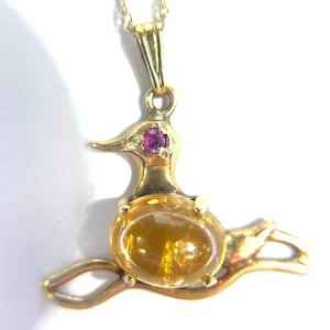 Puede incluir: Un collar con un colgante de oro con un diseño de pájaro con una piedra preciosa amarilla y un ojo de rubí rosa.