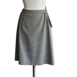 May include: A knee-length wrap skirt featuring a classic black and white houndstooth pattern. The skirt has a fitted waistband with a side tie closure, a straight silhouette, and a clean hemline, displayed on a mannequin for clear visibility.