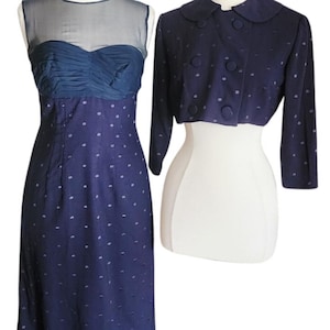 May include: A vintage navy blue dress and bolero jacket set. The dress has a sheer neckline and a ruched bodice. The dress and jacket are decorated with small white polka dots. The bolero jacket has a rounded collar and button fastenings.