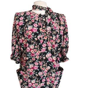 May include: A black dress featuring a vibrant floral pattern of pink, red, and yellow roses with green leaves. The dress has a round neckline, a front button closure, short sleeves with ruffled cuffs, two large front pockets, and a matching neck tie.