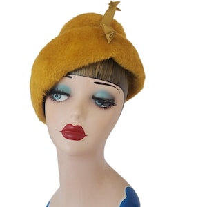 Puede incluir: Boina de color amarillo mostaza con un pequeño detalle decorativo. La boina está hecha de un material suave y texturizado y se muestra en una cabeza de maniquí. El maniquí tiene sombra de ojos azul y lápiz labial rojo. Un sombrero de estilo vintage.