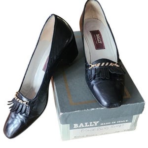 Può includere: Un paio di décolleté Bally nere con tacco basso e una catena dorata decorativa e frange. Le scarpe sono esposte accanto alla loro scatola originale, con il logo e il testo Bally.