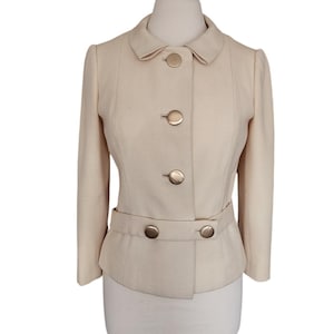 May include: A cream-colored jacket featuring a rounded collar, long sleeves, and a front button closure with four large, circular, metallic gold buttons. A horizontal belt detail at the waist is secured with two additional matching gold buttons.