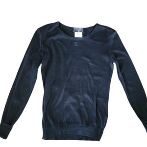 May include: A navy blue long-sleeved sweater with a crew neck. The sweater has a small, embroidered logo near the neckline and a label with the brand name. The cuffs and hem are ribbed.