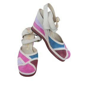 May include: A pair of wedge sandals featuring a multi-colored geometric suede pattern in shades of pink, purple, blue, and burgundy. The shoes have white leather ankle straps with gold-tone metal buckles and a white leather upper construction.