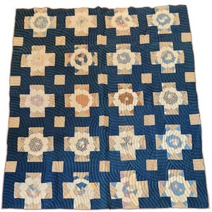 May include: A handmade quilt with a dark blue background and a repeating pattern of cross-shaped blocks. The blocks are made of various patterned fabrics in neutral tones, with a quilted design. The quilt is a square shape.