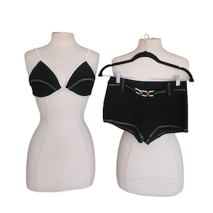 May include: A black bikini top with white trim and a pair of black shorts with a gold-colored buckle belt. The shorts are hanging on a black hanger. Both items have white stitching.