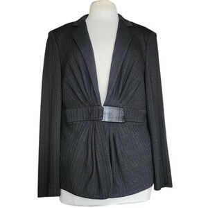 May include: A dark gray blazer featuring a subtle herringbone pattern, long sleeves, and a notched lapel. The front is gathered at the waist with a rectangular metallic buckle closure, creating a pleated effect above and below the waistline.