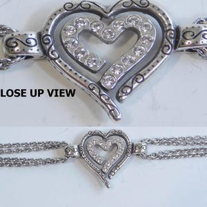 May include: Silver-toned bracelet featuring a heart-shaped pendant with a smaller heart inside, embellished with clear crystals. The bracelet has multiple delicate chain strands and a clasp closure. The words "CLOSE UP VIEW" are in the image.