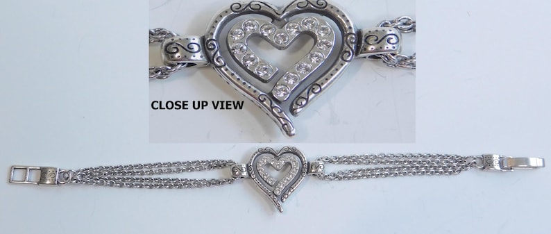 May include: Silver-toned bracelet featuring a heart-shaped pendant with a smaller heart inside, embellished with clear crystals. The bracelet has multiple delicate chain strands and a clasp closure. The words "CLOSE UP VIEW" are in the image.